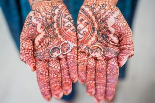 Intricate henna tattoo design on a pair of hands holding silver and gold wedding and engagement rings