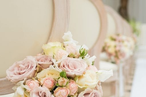 Pastel pink and white rose bouquets attached to the back of a white oval ceremony chairs in the Orangery at Blenheim Palace