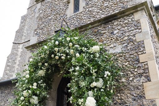 A stunning white and green floral arch at the entrance of a countryside church