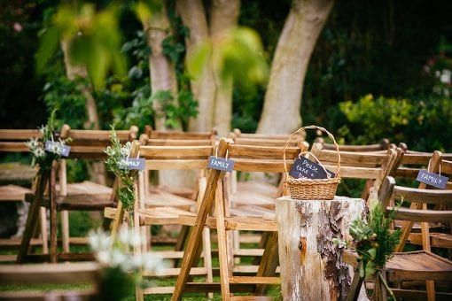 Rustic wedding ceremony set in a rose garden on a private estate with wooden chairs styled with green foliage