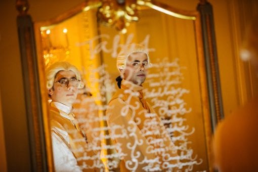 Waiters styled for a Marie Antoinette themed party in front of a seating plan with guest names calligraphed onto a gold mirror